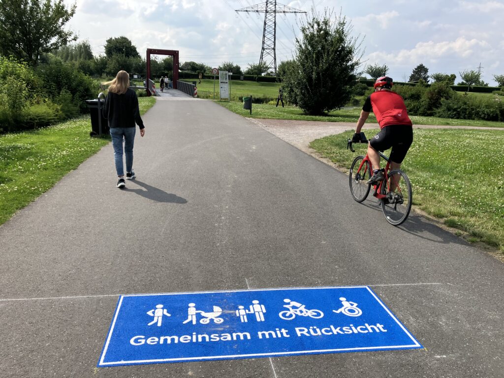 Im Vordergrund ein blaues Piktogramm "Gemeinsam mit Rücksicht" mit verschiedenen Symbolen; dahinter links eine Fußgängerin und rechts ein Radfahrer; im Hintergrund eine Brücke