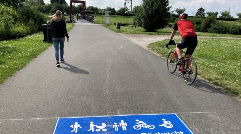 Im Vordergrund ein blaues Piktogramm "Gemeinsam mit Rücksicht" mit verschiedenen Symbolen; dahinter links eine Fußgängerin und rechts ein Radfahrer; im Hintergrund eine Brücke