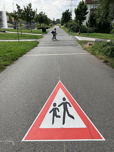 Im Vordergrund ein markiertes Verkehrszeichen 136 "Kinder"; Weg geht geradeaus.