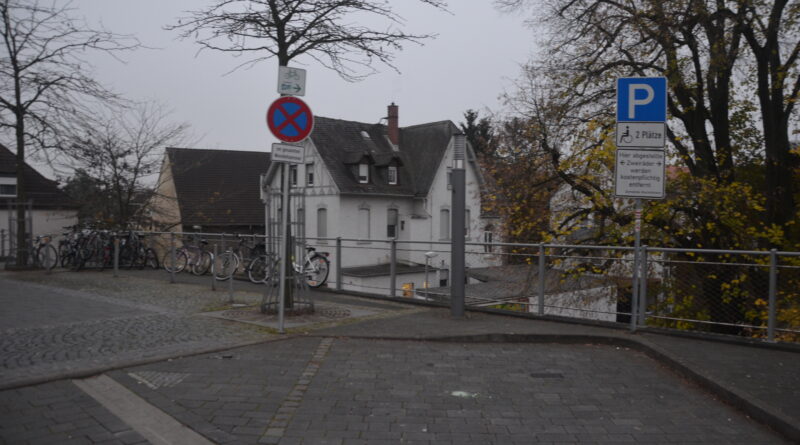 im Vordergrund ein Parkplatz, als s. g. Behinderten-Stellplatz beschildert; ein HBR-Wegweiser nach rechts zu einem Gerländer