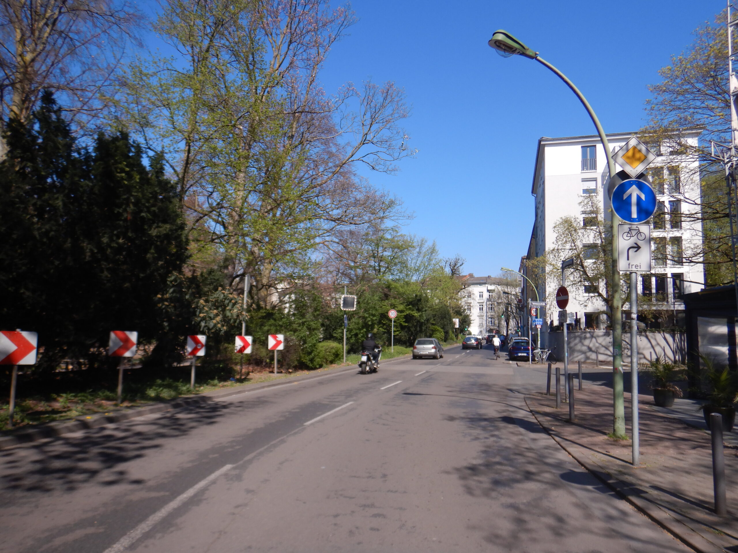 Rechts steht "Vorgeschriebene Fahrtrichtung geradeaus" als Verkehrszeichen neben der Fahrbahn, darunter ein Zusatzzeichen mit dem Sinnbild Fahrrad, einem Pfeil nach rechts und dem Wort frei.