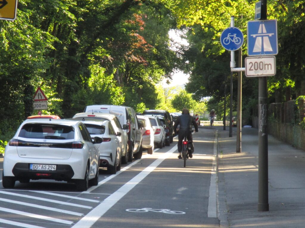 Links parken Autos, in der Mitte der Radfahrstreifen. Auf ihzm fährt ein Radfahrer vom Fotostandpunkt weg.