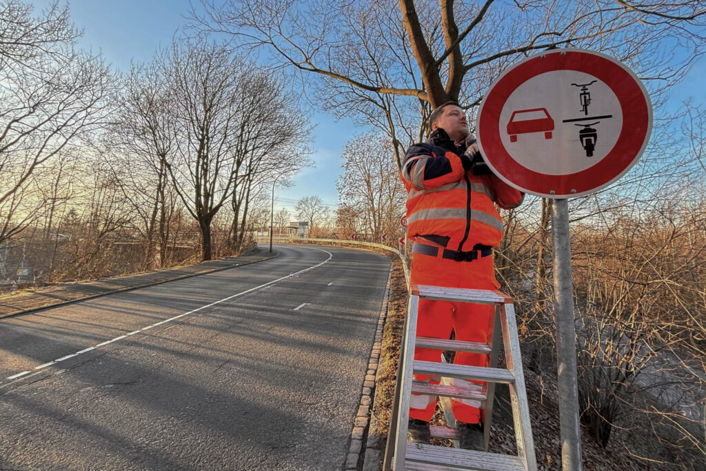 Die Straße biegt im Hintergrund nach links ab. An den Seiten stehen unbelaubte Bäume. Rechts vorne montiert ein Mitarbeiter der Stadt das neue Verkehrszeichen. Er steht auf einer Leiter.