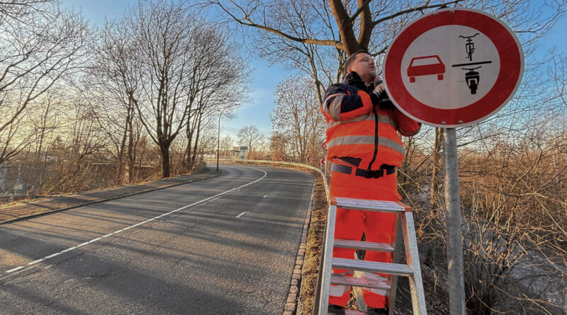 Die Straße biegt im Hintergrund nach links ab. An den Seiten stehen unbelaubte Bäume. Rechts vorne montiert ein Mitarbeiter der Stadt das neue Verkehrszeichen. Er steht auf einer Leiter.