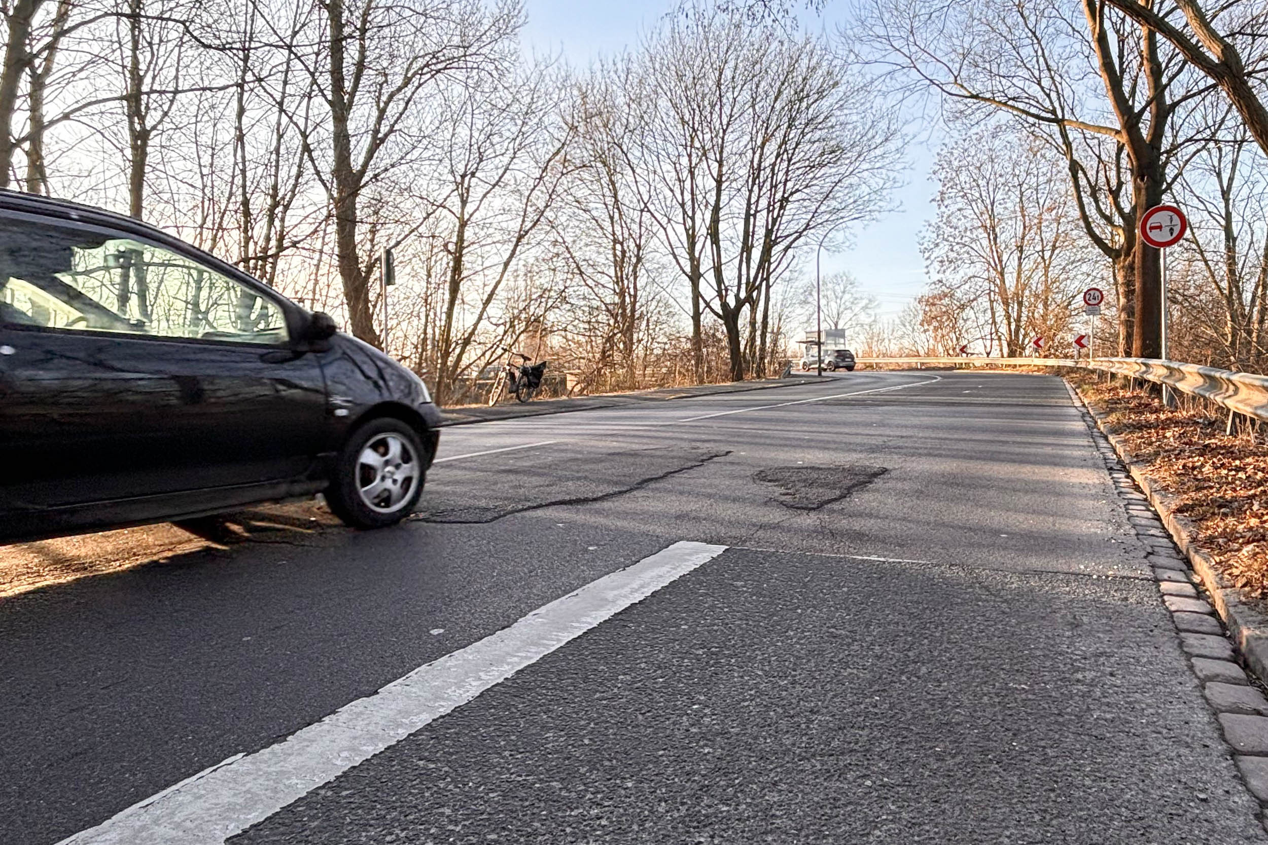 Die Straße biegt im Hintergrund nach links ab. An den Seiten stehen unbelaubte Bäume. In etwas Entfernung steht das neue Schild. Links fährt ein Auto in Blickrichtung.