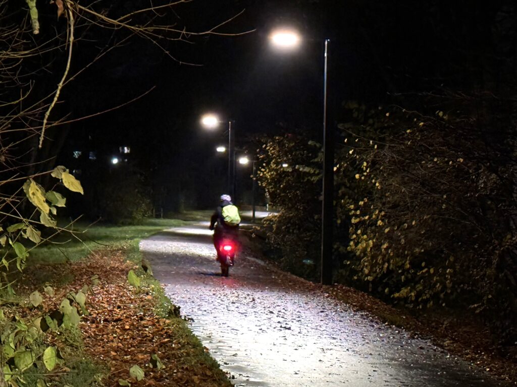 Der Weg macht eine leichte Kurve nach rechts. Auf der rechten Seite stehen Straßenlaternen, die leuchten. Der Himmel ist dunkel. Auf dem Weg fährt jemand mit dem Rad in Blickrichtung.